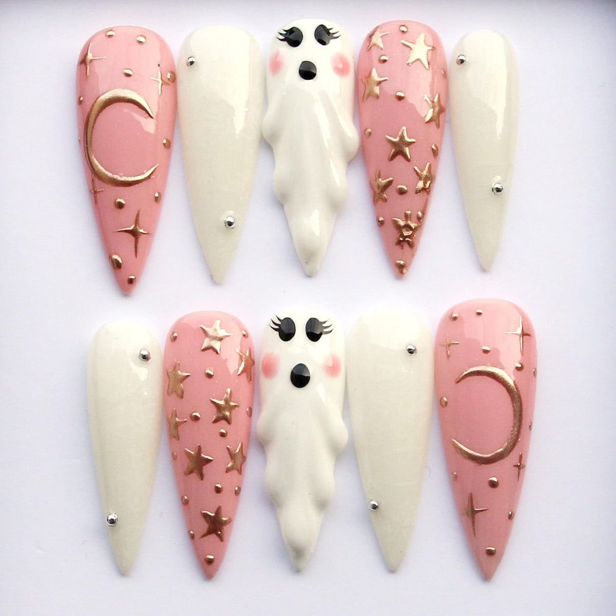 A set of ten long, almond-shaped false nails. The nails feature a mix of designs, including solid white, pale pink with gold stars and moons, and ghost-shaped accents. Each nail has a glossy finish.