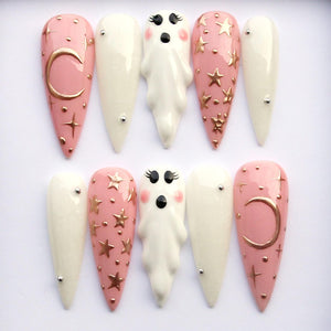 A set of ten long, almond-shaped false nails. The nails feature a mix of designs, including solid white, pale pink with gold stars and moons, and ghost-shaped accents. Each nail has a glossy finish.