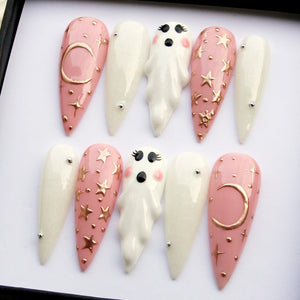 A set of ten long, almond-shaped artificial nails. The nails feature a variety of designs, including a pink base with gold stars and moons, white nails with silver accents, and 3D ghost figures. The nails are arranged in a row.