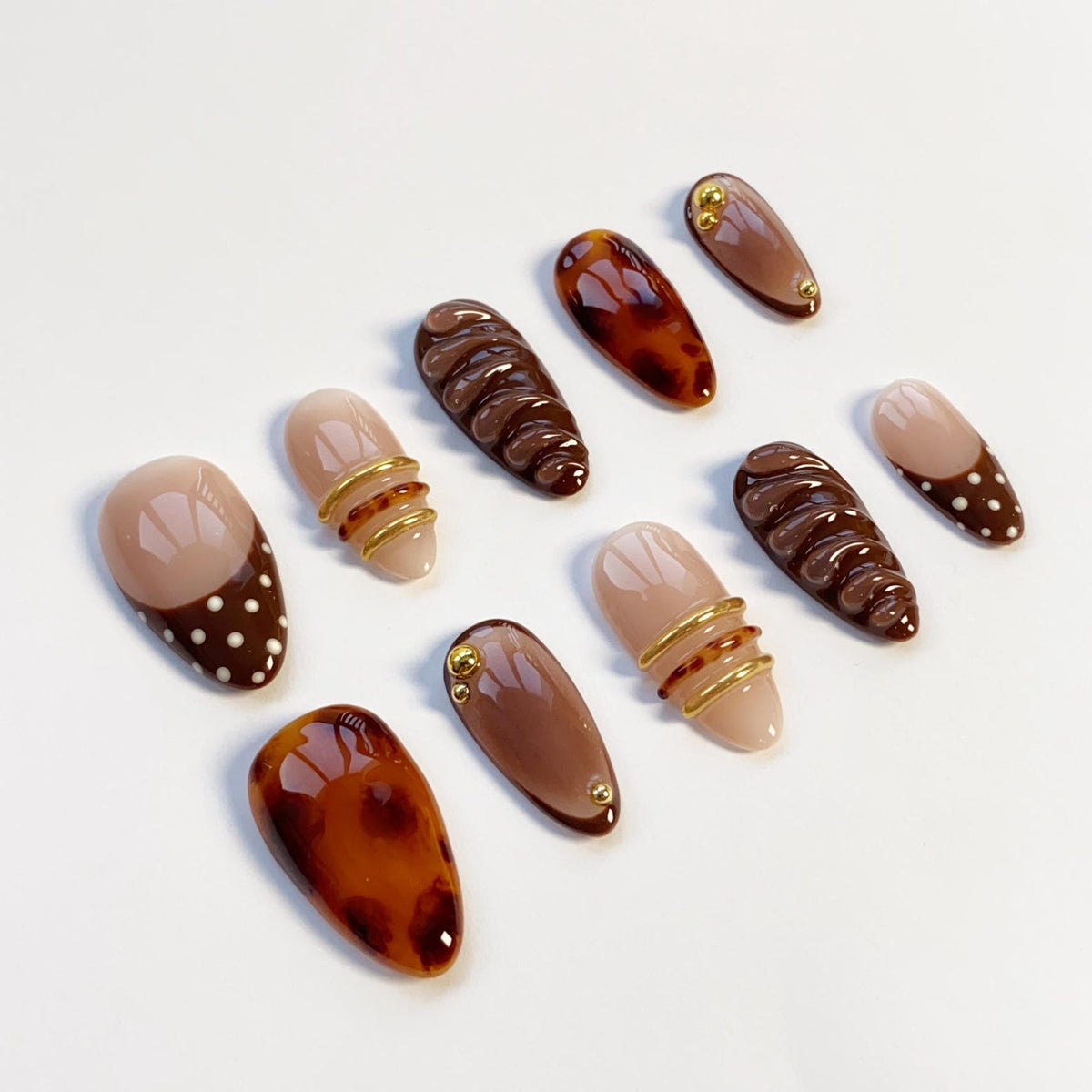 A collection of artificial nails in various designs. Some nails feature a tortoiseshell pattern, while others have a solid brown or nude color. Several nails are adorned with gold-colored accents and some have a polka dot pattern.