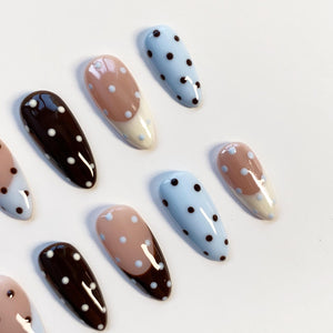 A collection of almond-shaped false nails. The nails feature various designs, including light blue with black polka dots, brown with white polka dots, and nude with white polka dots and a white tip.
