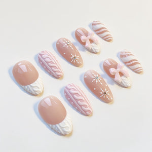 A collection of artificial nails in various designs. Some nails are light pink with white snowflake accents, others feature a cable knit pattern, and some have a bow design. The nails are oval-shaped and have a glossy finish.