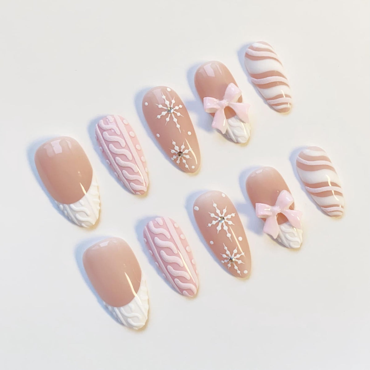 A set of ten almond-shaped false nails. The nails feature a variety of designs, including a light pink base with white snowflake accents, pink bows, and white cable knit patterns. Some nails have a nude base with a white tip.