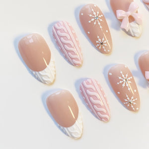 A collection of almond-shaped artificial nails in various designs. Some nails feature a light pink base with white snowflake accents, while others have a cable knit pattern or a French tip design with a white textured edge.