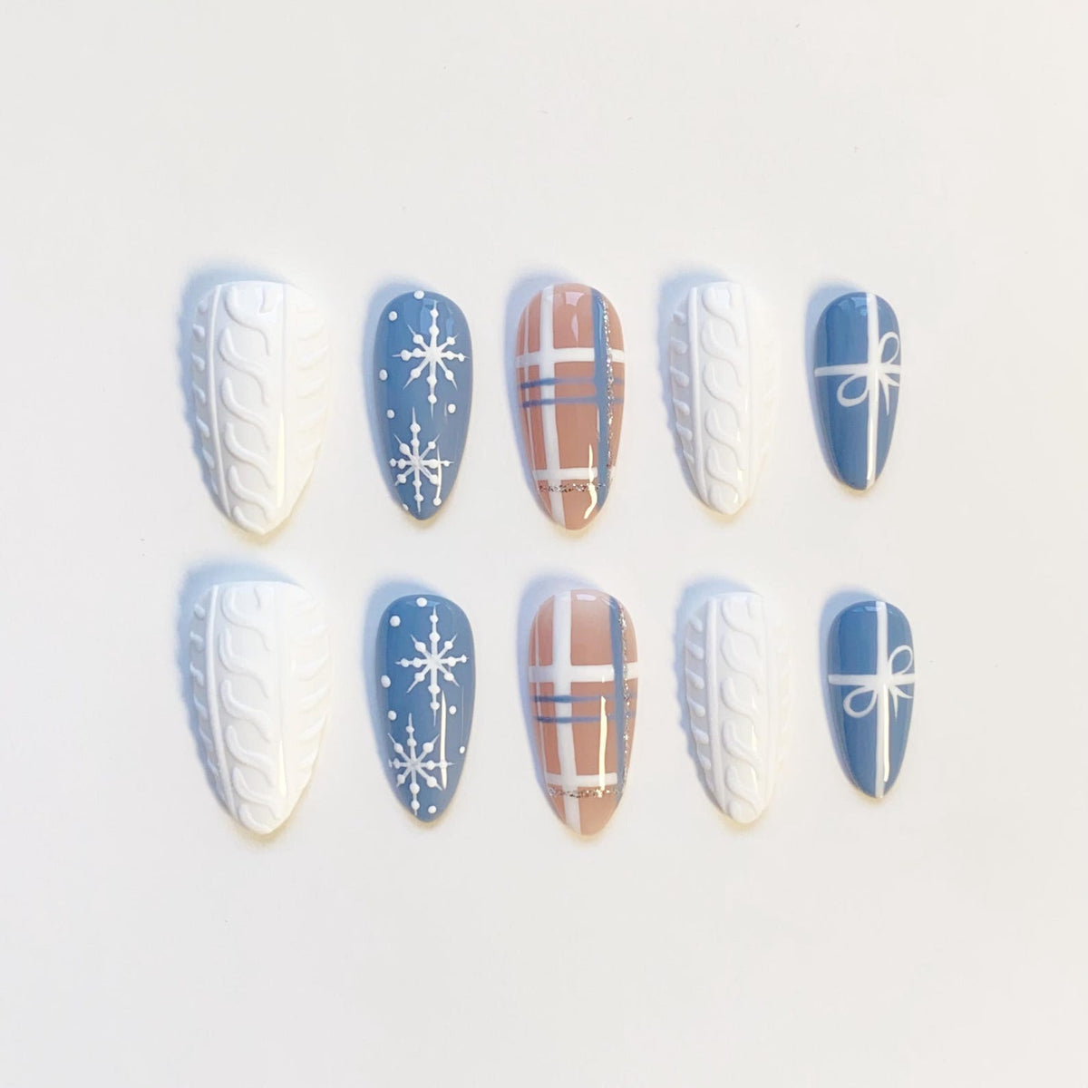 A set of ten almond-shaped false nails. Designs include white cable knit, blue with white snowflakes, plaid, and blue with a white bow. The nails are arranged on a white surface.