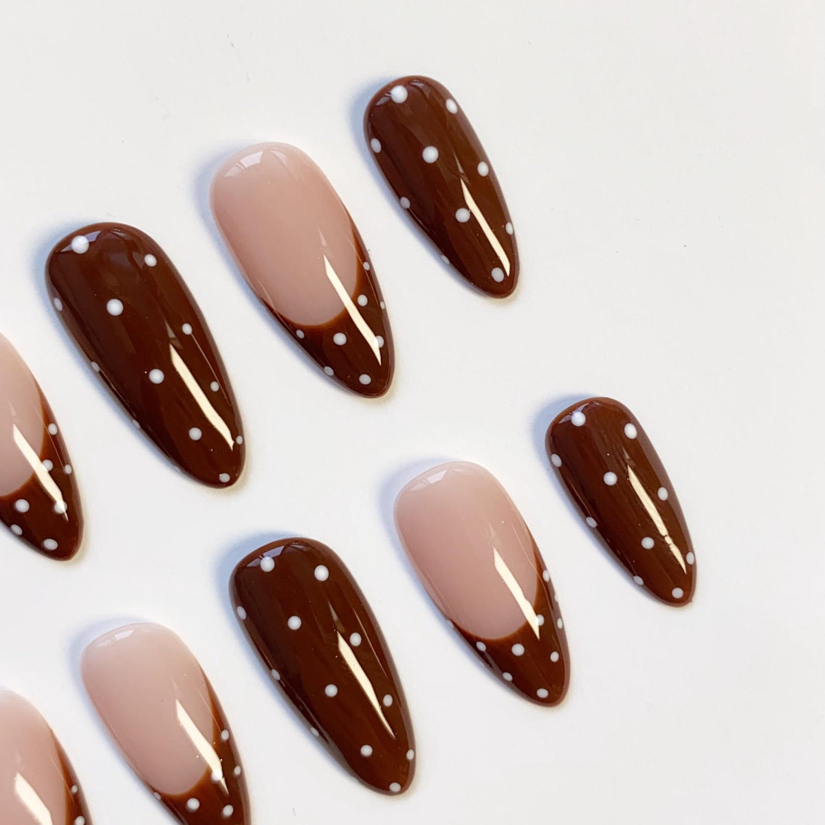 A set of artificial nails with a brown and white polka dot design. Some nails have a nude base with a brown French tip. The nails are oval-shaped and are arranged on a white surface.