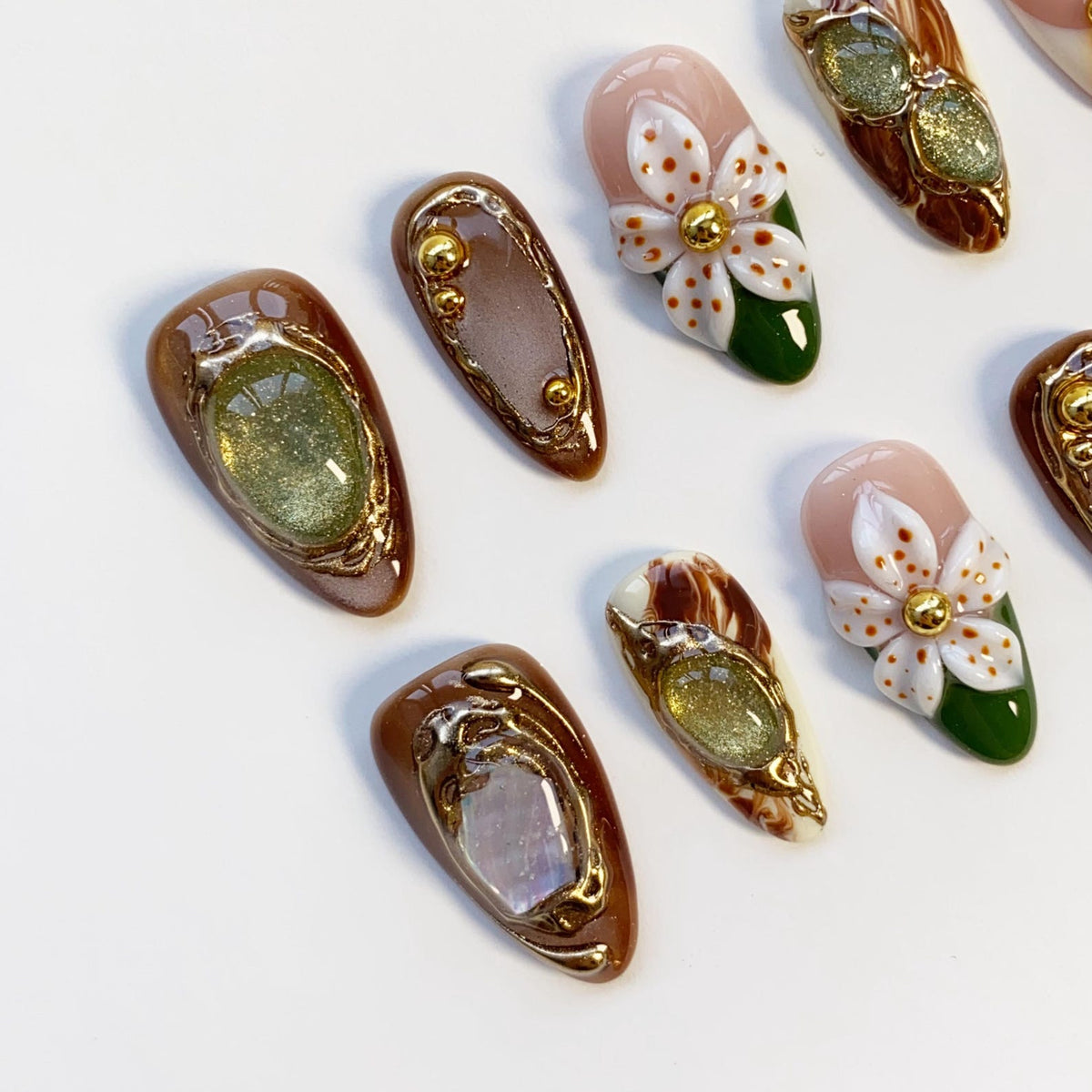 A collection of artificial nails with various designs. Some nails feature a brown base with gold-colored embellishments and glittery accents. Others showcase a floral design with white petals, gold centers, and green leaves. The nails are arranged on