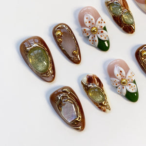 A collection of artificial nails with various designs. Some nails feature a brown base with gold-colored embellishments and glittery accents. Others showcase a floral design with white petals, gold centers, and green leaves. The nails are arranged on