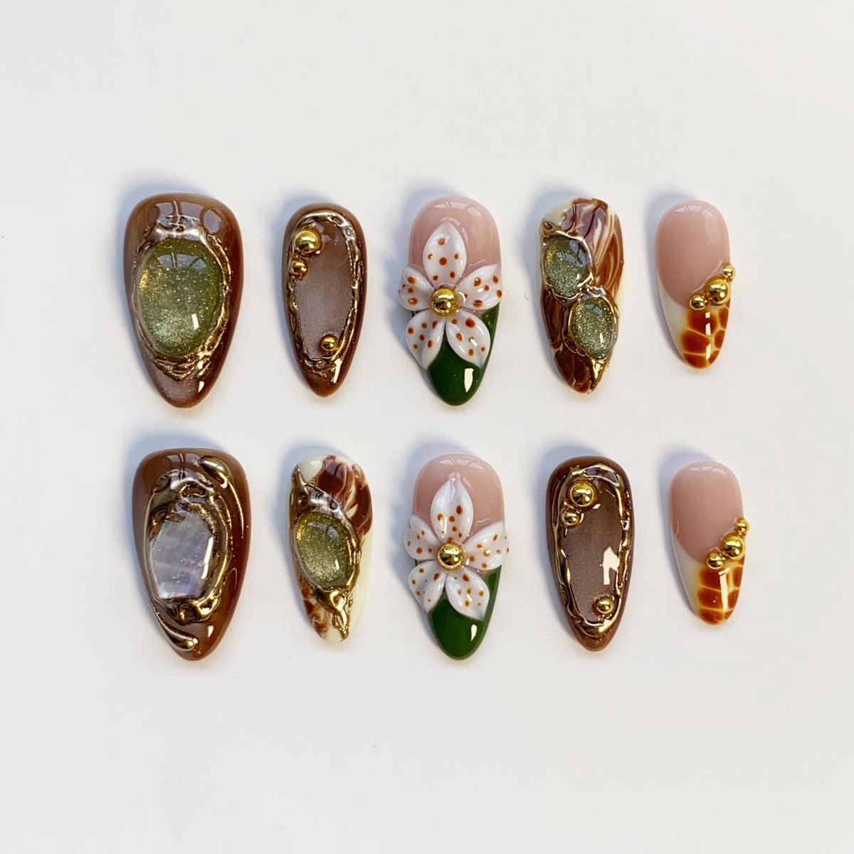 A set of ten artificial nails with various designs. Some nails are brown with gold accents and embedded gems. Others feature white floral designs with green bases. A few have a nude base with gold embellishments.