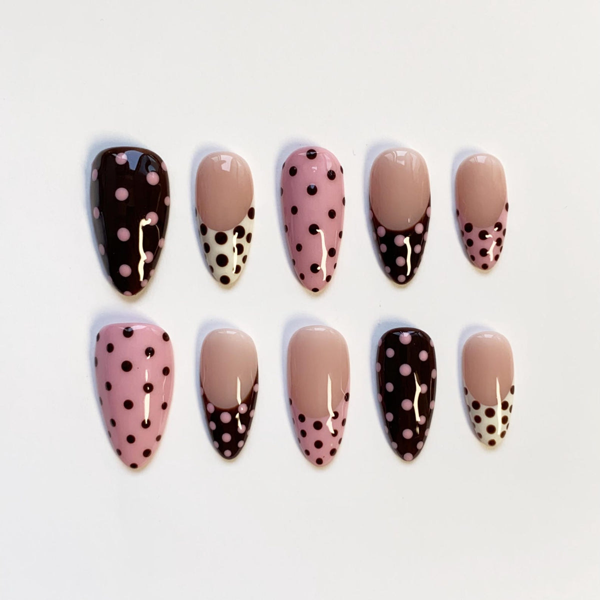 A set of ten almond-shaped false nails. The nails feature a variety of designs, including solid colors, pink with black polka dots, and a French tip design with black and white polka dots.