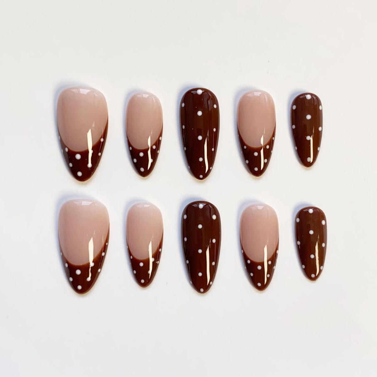 A set of ten almond-shaped false nails. The nails feature a light beige base with a dark brown French tip adorned with small white polka dots. The design is consistent across all nails, offering a cohesive look.