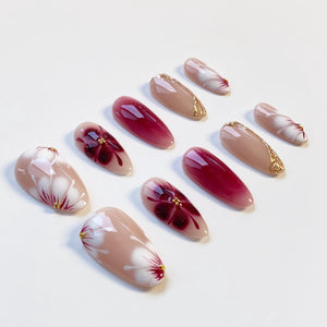 A collection of almond-shaped artificial nails. The nails feature various designs, including floral patterns in white and red, solid burgundy, and nude with gold accents. The nails are arranged on a white surface.