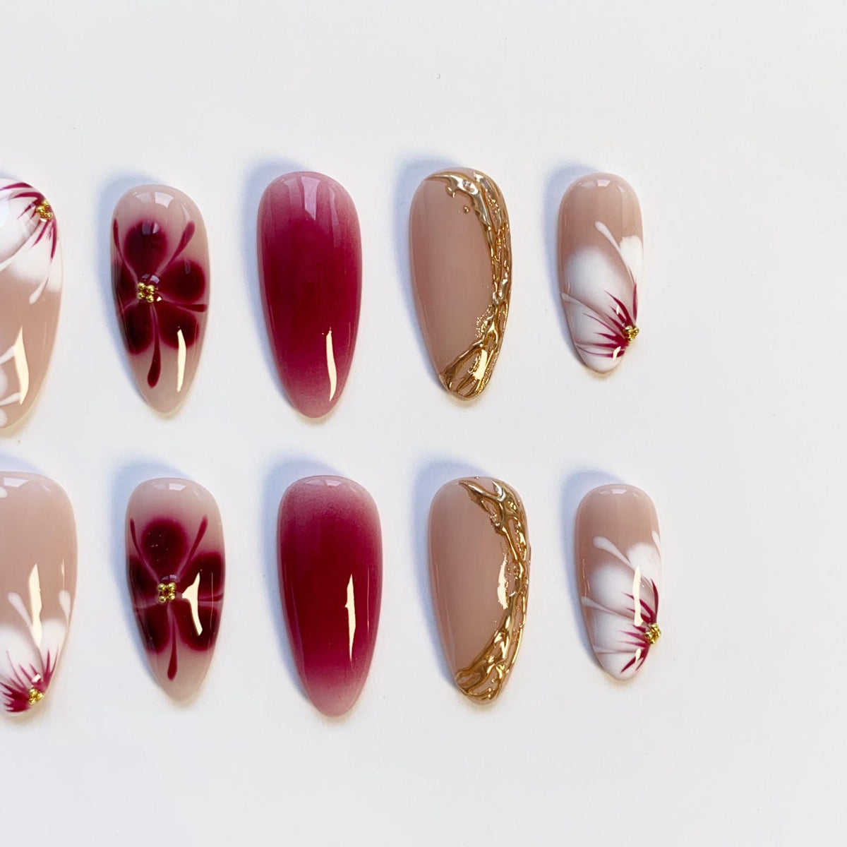 A set of artificial nails in various designs. Some nails are a solid deep red, others feature a floral design with red and gold accents. Several nails have a gold decorative edge. The nails are oval-shaped and set against a white background.