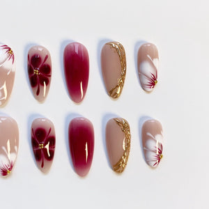 A set of artificial nails in various designs. Some nails are a solid deep red, others feature a floral design with red and gold accents. Several nails have a gold decorative edge. The nails are oval-shaped and set against a white background.