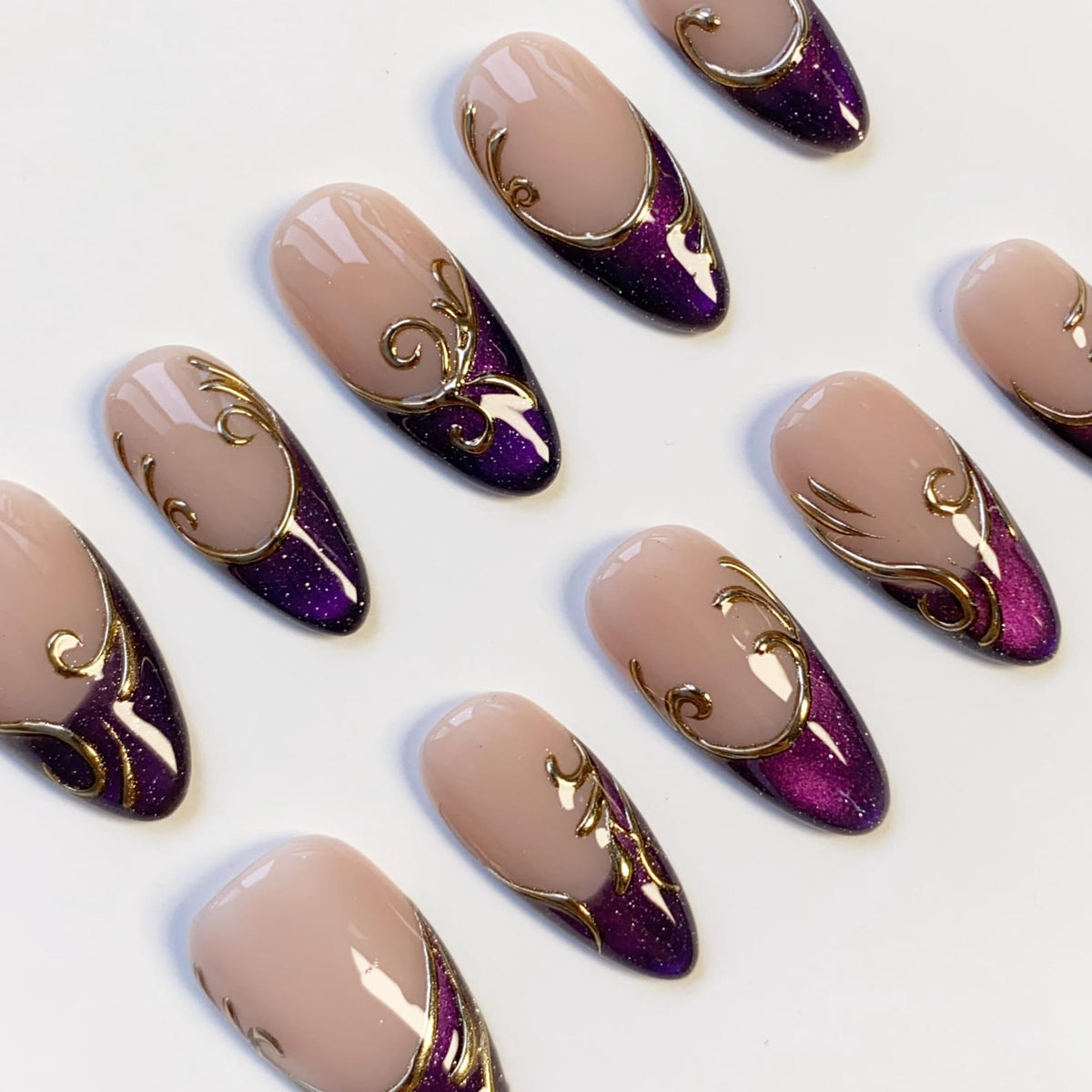 A collection of artificial nails with a light pink base, a deep purple glittered lower section, and gold filigree accents. The nails are oval-shaped and arranged on a white surface, showcasing a glamorous design.
