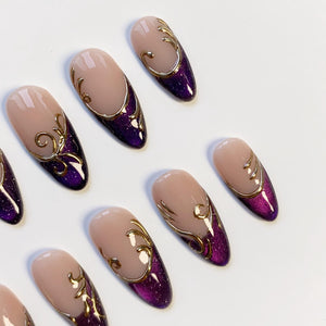 Oval-shaped false nails with a nude base and a deep purple glitter gradient. Each nail features gold filigree designs along the edges. The nails are arranged on a white surface, showcasing a glamorous nail art style.