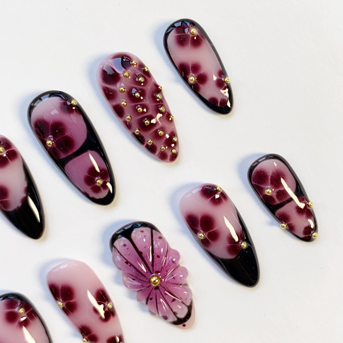 A collection of artificial nails with a floral design. The nails feature a gradient of pink to dark purple, with gold accents. Some nails have a 3D flower design, while others have gold studs.