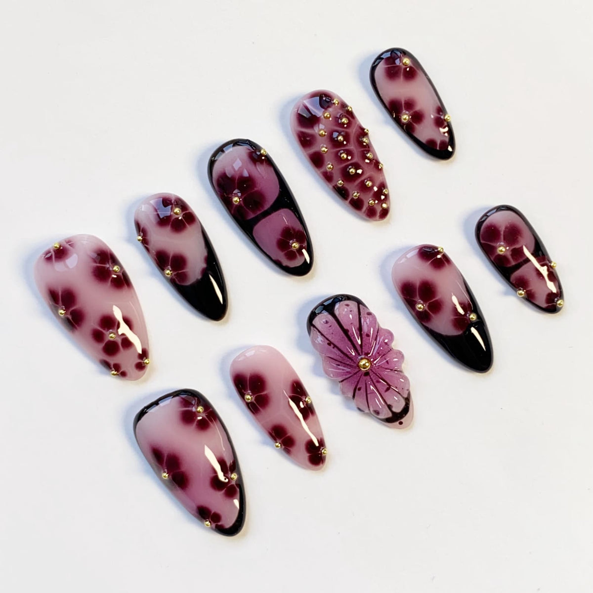 A collection of artificial nails with various designs. The nails feature a gradient of pink and purple hues, adorned with floral patterns and small gold embellishments. Some nails have a black base, while others showcase a 3D flower design.