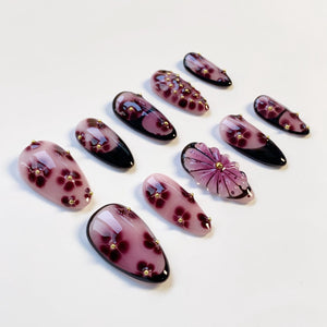 A collection of artificial nails in various designs. The nails are primarily shades of purple and pink, with floral and geometric patterns. Some nails feature small gold accents.