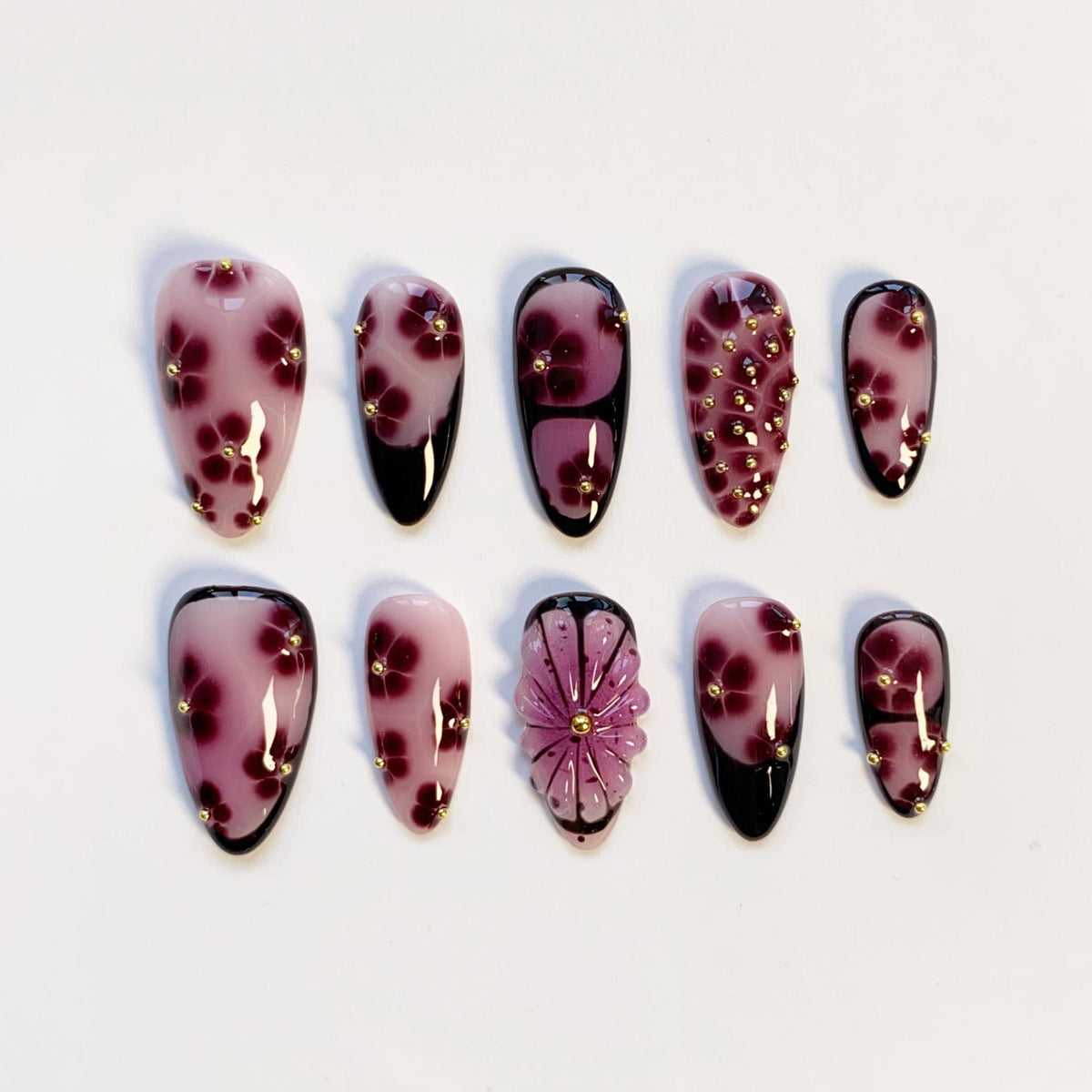 Set of ten artificial nails in various designs. The nails feature a light pink base with dark red floral patterns and gold accents. Some nails have black tips, and one has a 3D flower design.