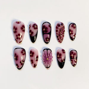 Set of ten artificial nails in various designs. The nails feature a light pink base with dark red floral patterns and gold accents. Some nails have black tips, and one has a 3D flower design.