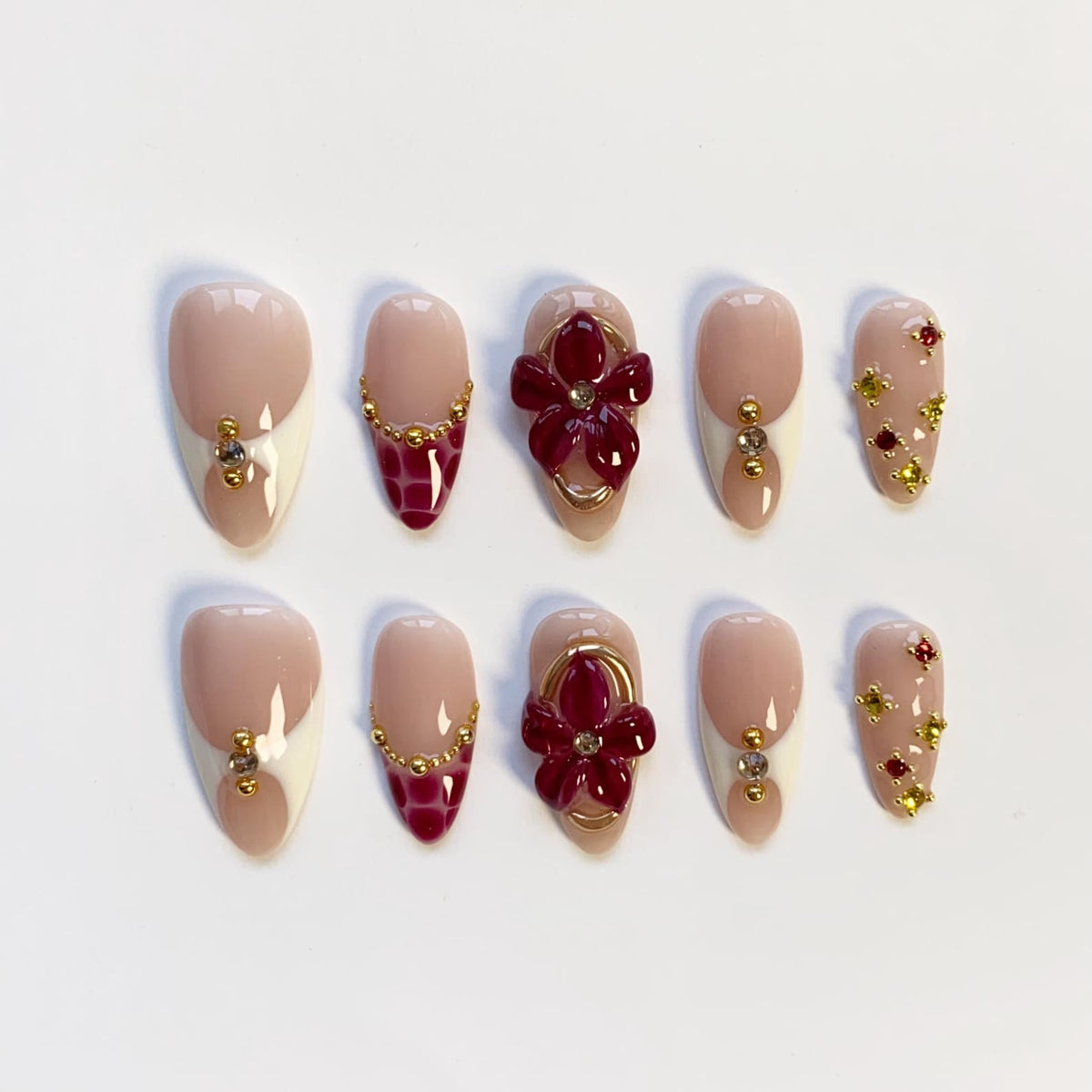 A set of ten artificial nails in various designs. Some nails feature a nude base with a white tip and gold accents. Others showcase a burgundy crocodile pattern or a 3D burgundy flower design with gold trim. Some nails have gold and jewel accents.