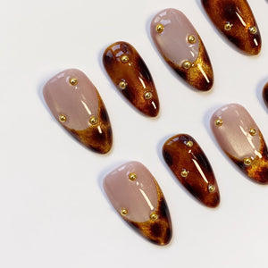 Oval-shaped artificial nails in a variety of designs. Some nails are a light mauve color with gold accents and gold studs. Other nails have a brown and gold tortoiseshell pattern with gold studs.