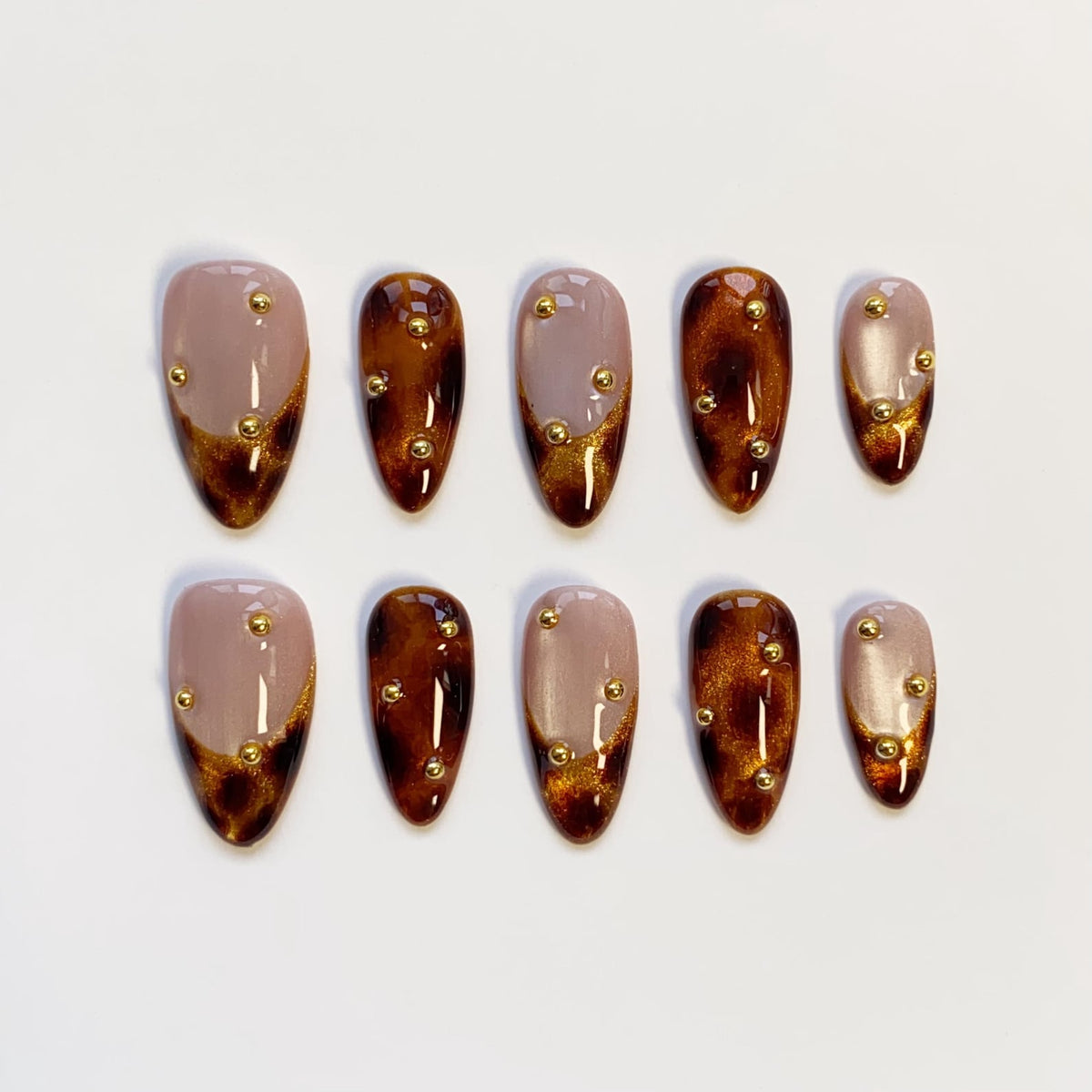 A set of ten almond-shaped press-on nails. Each nail features a gradient design, transitioning from a nude base to a dark brown tip with gold glitter accents. Small gold studs are evenly spaced across each nail.