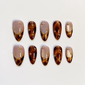 A set of ten almond-shaped press-on nails. Each nail features a gradient design, transitioning from a nude base to a dark brown tip with gold glitter accents. Small gold studs are evenly spaced across each nail.