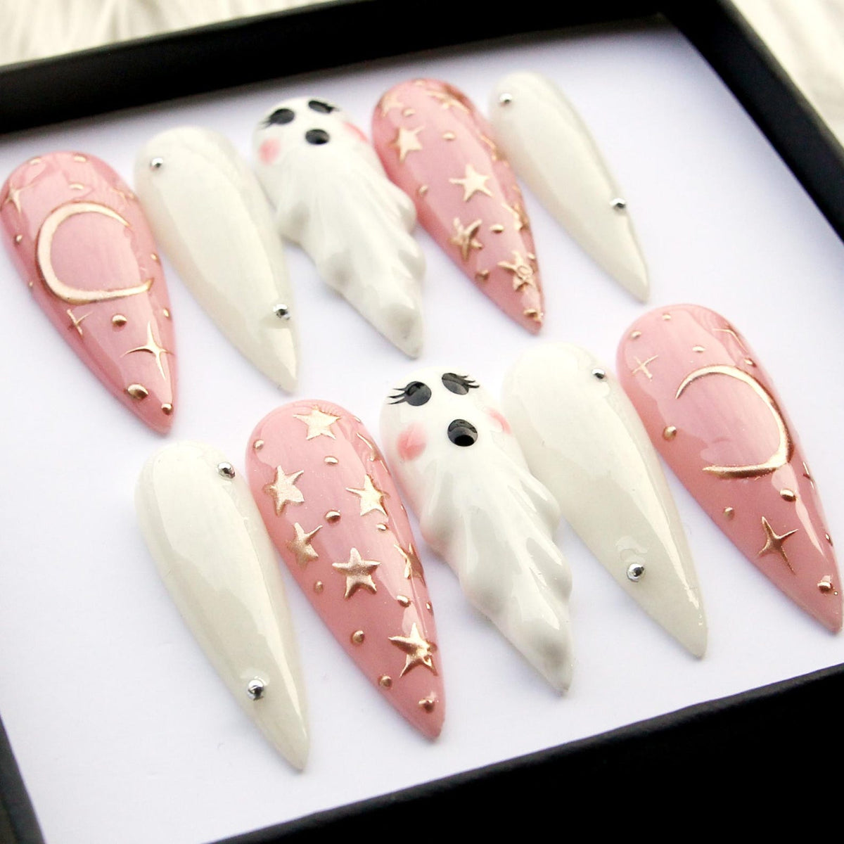 A set of ten long, pointed false nails. The nails feature a variety of designs, including white, pale pink with gold stars, and pink with a gold crescent moon. Some nails have small ghost figures.