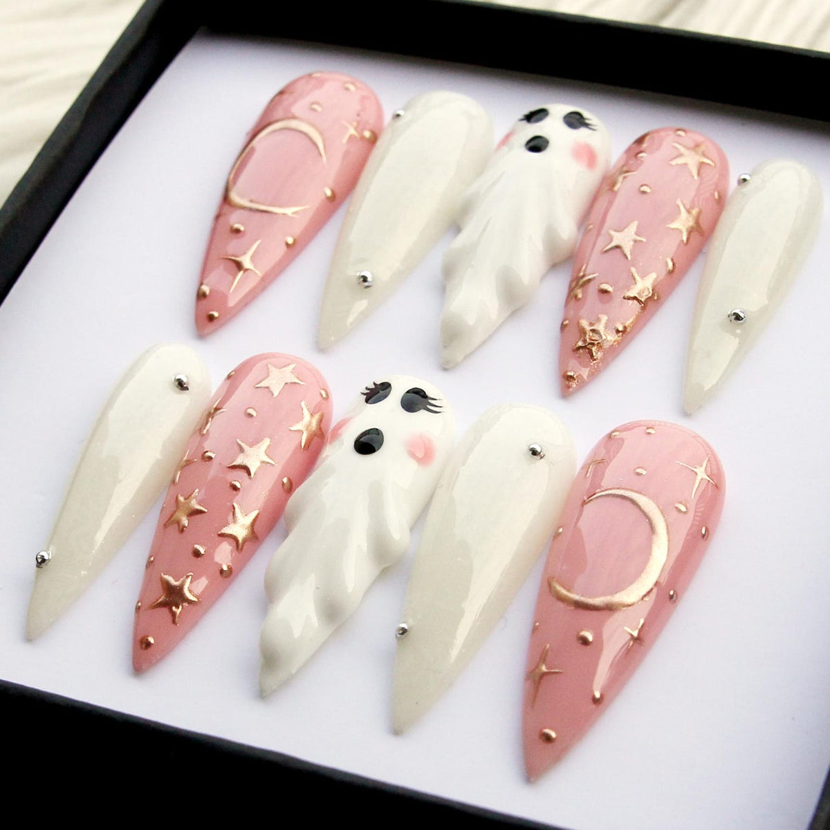 A set of ten long, almond-shaped false nails. The nails feature a mix of designs, including pale pink with gold stars and moons, white with silver accents, and 3D ghost figures. The nails are arranged in a black box.