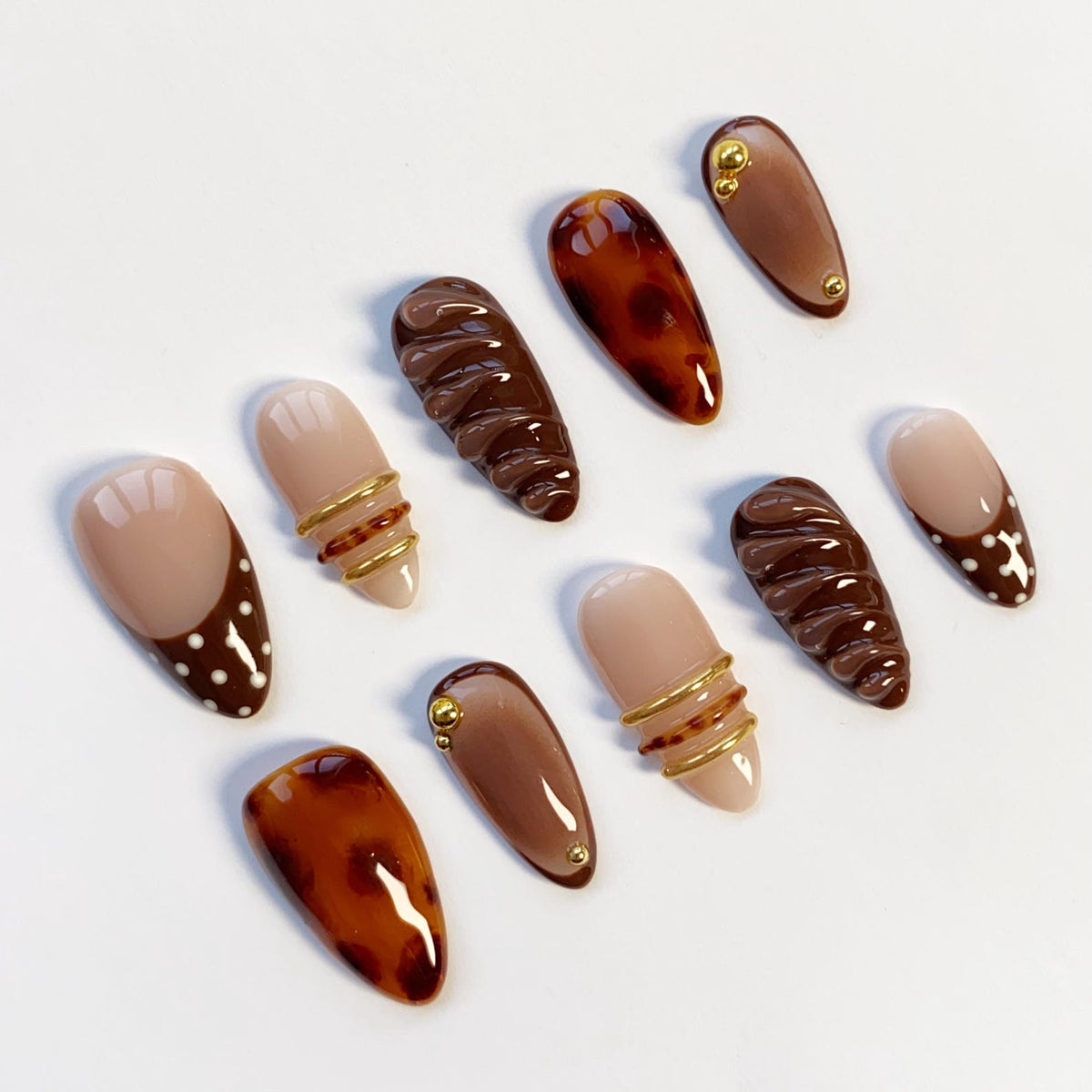 A collection of ten almond-shaped artificial nails in various designs. Some nails are solid brown, others feature tortoiseshell patterns, and some have gold wire accents. Several nails have a nude base with brown polka dots or a brown tip.