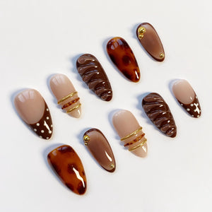 A collection of ten almond-shaped artificial nails in various designs. Some nails are solid brown, others feature tortoiseshell patterns, and some have gold wire accents. Several nails have a nude base with brown polka dots or a brown tip.