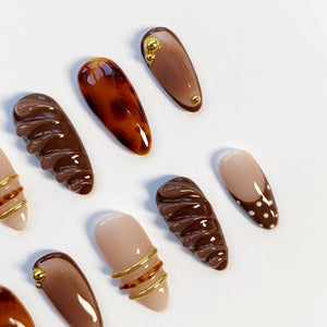 A collection of artificial nails in various designs and colors. Some nails are solid brown, others feature gold accents, and one has a tortoiseshell pattern. One nail has a white tip with brown polka dots. The nails are arranged on a white surface.