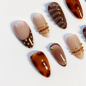 A collection of artificial nails in various designs. Some nails are solid colors, while others feature patterns like polka dots, swirls, and gold accents. The color palette includes shades of brown, beige, and gold.
