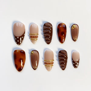 A set of ten artificial nails in various designs. The nails feature a mix of solid colors, tortoiseshell patterns, and decorative elements like gold rings and dots. The color palette includes shades of brown, beige, and gold.