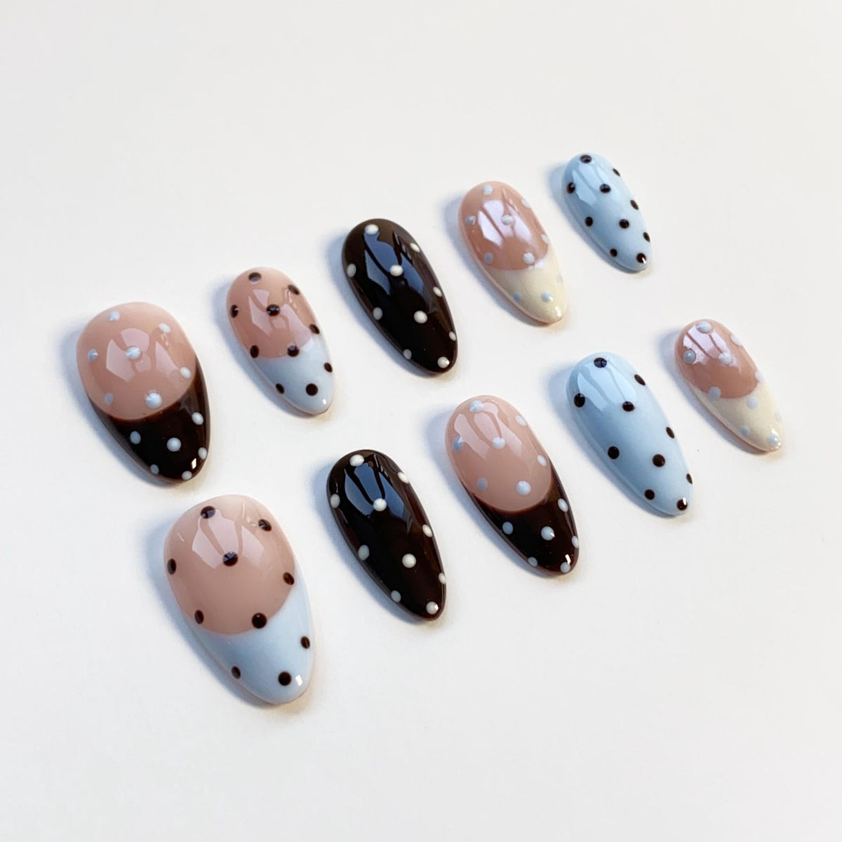 A set of ten artificial nails in various designs. Some nails are black with white polka dots, others are light blue with white polka dots, and some have a two-tone design of brown and light blue with white polka dots.