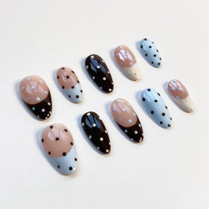 A set of ten artificial nails in various designs. Some nails are black with white polka dots, others are light blue with white polka dots, and some have a two-tone design of brown and light blue with white polka dots.