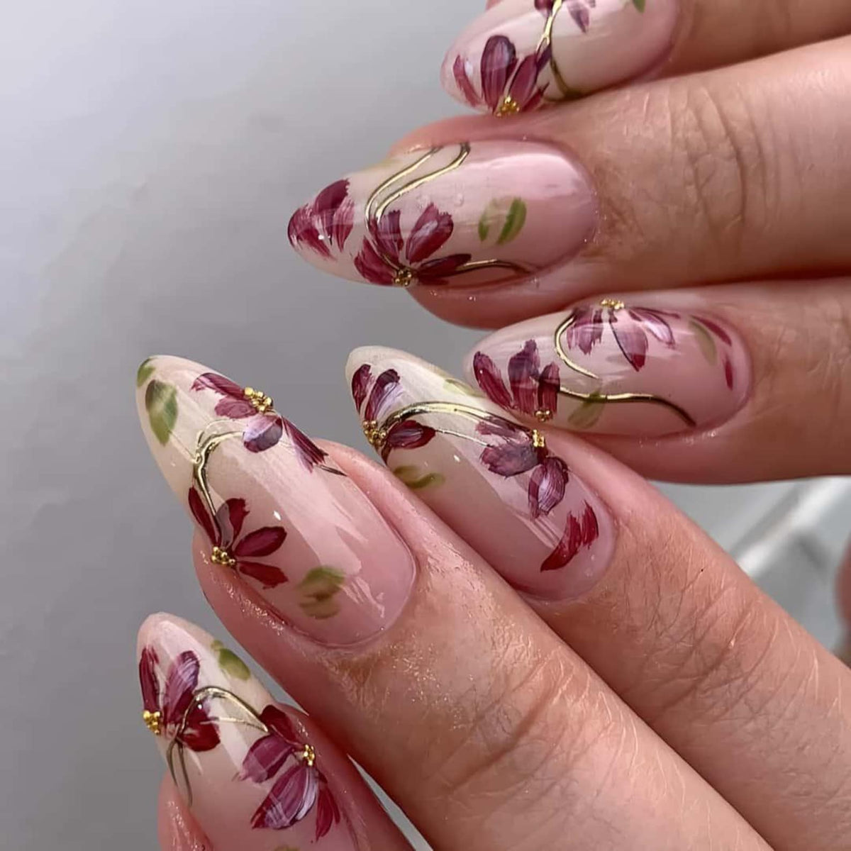 Almond-shaped nails with a floral design. The nails are painted with a light pink base and feature burgundy flowers with gold accents. Green leaves and small gold beads complete the design, creating a delicate and artistic look.