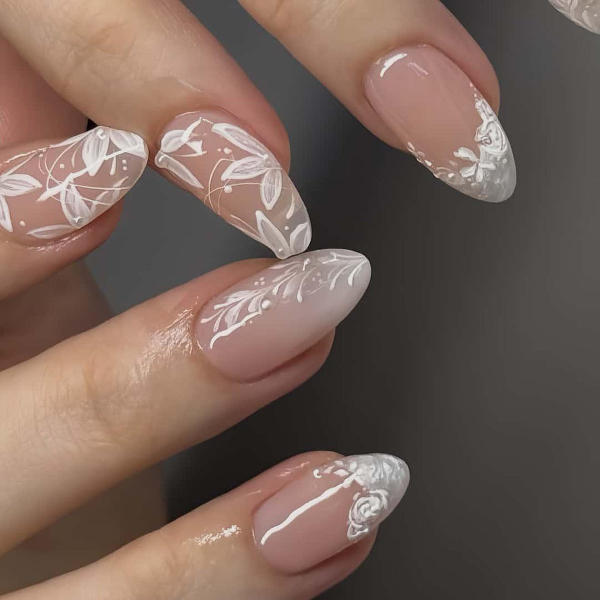 Close-up of manicured nails with a soft pink base and intricate white floral designs. The nails are oval-shaped and feature delicate, hand-painted patterns of leaves and flowers, creating an elegant and artistic look.
