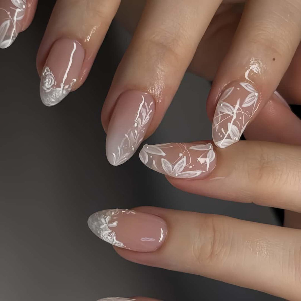Close-up of manicured nails with a natural pink base and white floral designs. The nails have an oval shape and feature delicate white patterns, including leaves and floral accents, creating an elegant and artistic look.