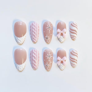 A set of ten almond-shaped false nails. The nails feature a variety of designs, including a light pink base with white cable knit accents, snowflake patterns, and pink bows. Some nails have a nude base with white cable knit tips.