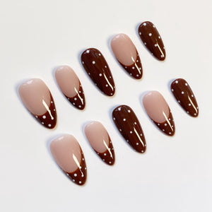 A set of almond-shaped false nails. The nails alternate between a solid dark brown color with white polka dots and a nude base with a dark brown tip and white polka dots.