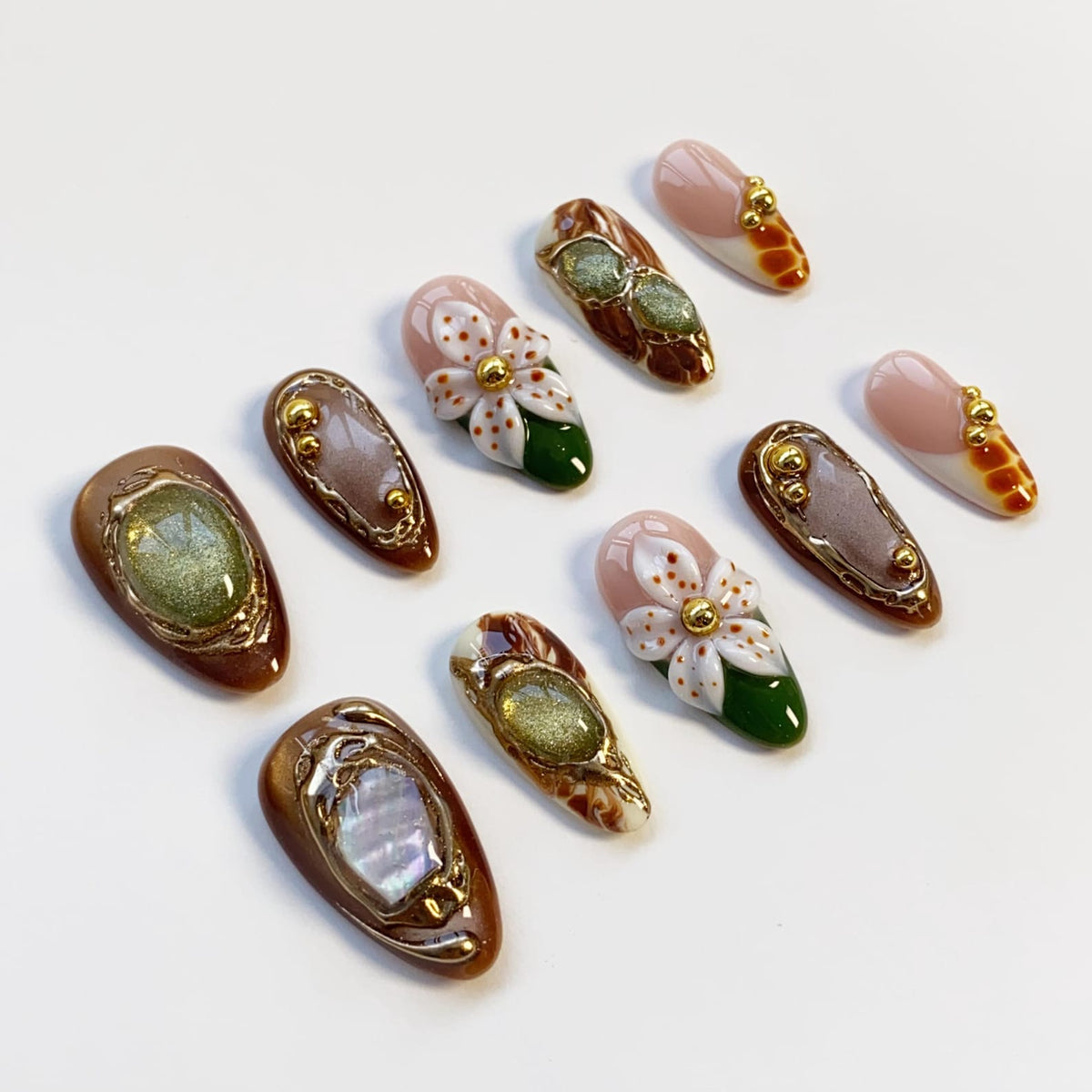 An assortment of almond-shaped, artificial nails with various designs. Some nails feature floral patterns with white petals and gold centers, while others have brown, gold, and iridescent accents. The nails are arranged on a white surface.