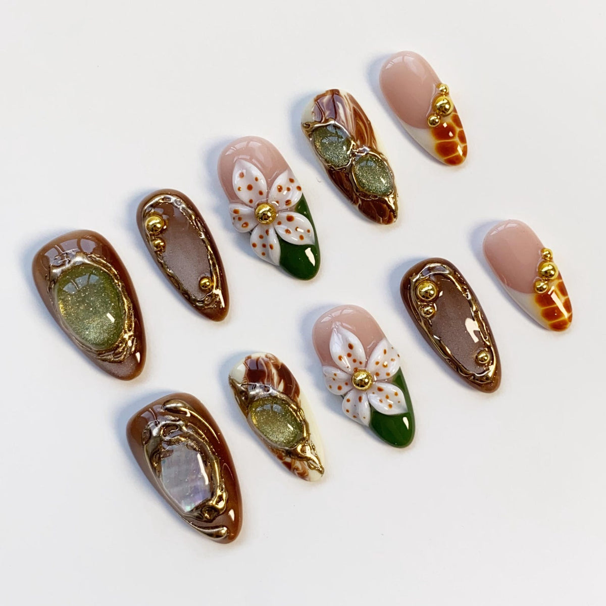 A collection of ten artificial nails with various designs. Some nails feature a light pink base with gold accents, while others have brown and gold frames with green or white floral designs. The nails are arranged on a white surface.