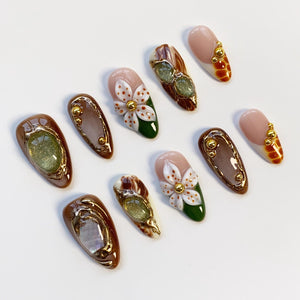 A collection of ten artificial nails with various designs. Some nails feature a light pink base with gold accents, while others have brown and gold frames with green or white floral designs. The nails are arranged on a white surface.