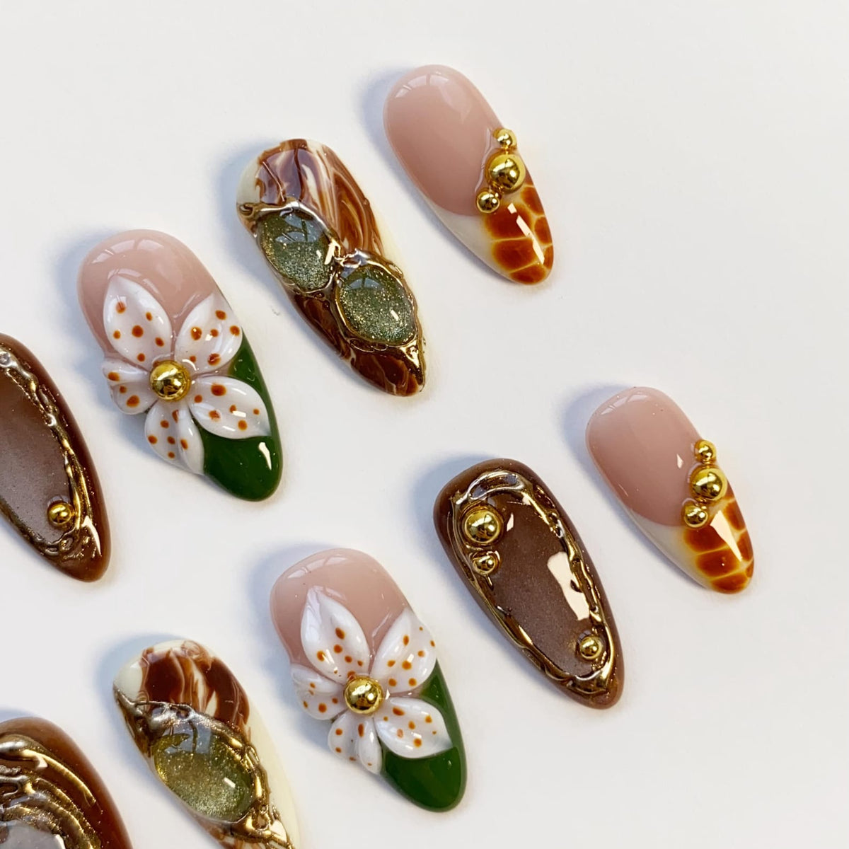 A collection of artificial nails with various designs. Some nails feature a nude base with gold accents, while others showcase intricate floral patterns with white petals and gold centers. Other nails have a brown marble effect.