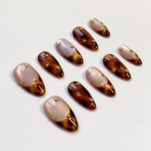 A set of ten artificial nails in various designs. Some nails are a light mauve color with gold accents, while others are a dark brown with gold flecks. Each nail has a rounded shape and is adorned with small gold studs.