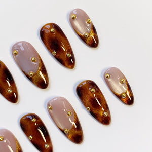 Oval-shaped press-on nails in a variety of designs. Some nails are a light mauve color with gold glitter accents, while others are a dark brown with gold glitter accents. Each nail has small gold studs.