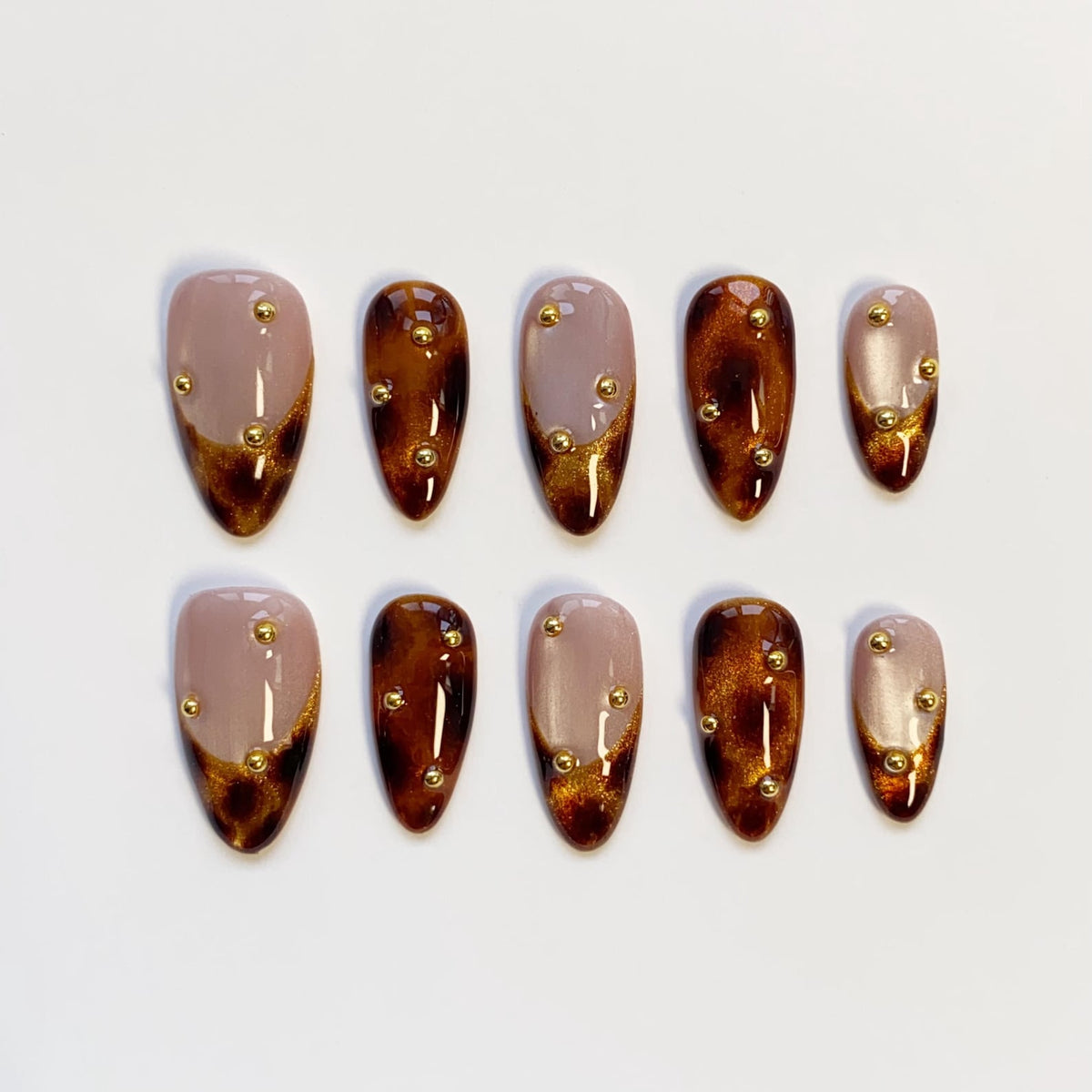 Set of ten almond-shaped false nails. Each nail features a gradient design with a nude base, transitioning to a dark brown tip with gold glitter accents. Small gold studs are evenly spaced across each nail.
